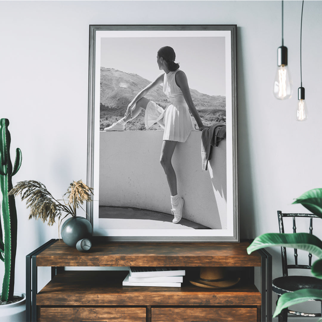 Fashion model in tennis outfit seated on wall looking at mountains by Toni Frissell poster artwork, framed and displayed in home decor